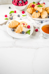 Sweet summer dessert, homemade baked mini croissants with berry jam, served with tea, fresh raspberries, blueberries and mint. On a white marble table, copy space