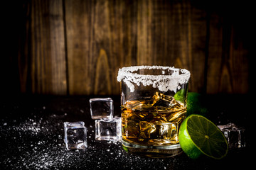 Two tequila shot glasses on dark background, with ice cubes, salt and limes copy space