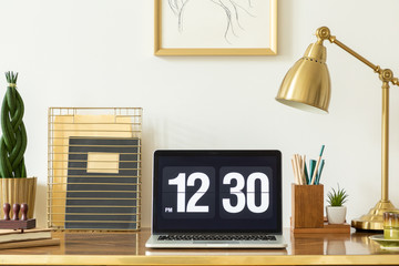 Laptop showing the time, notebooks, plant and lamp on a desk in an office interior. Real photo