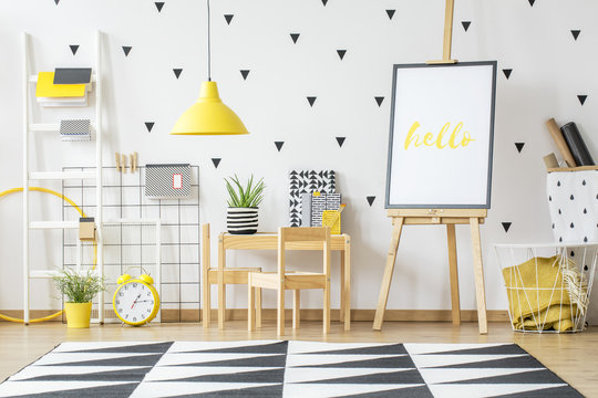 Poster on a wooden easel and a small desk for children in a white, scandi style preschool room interior with white wall and vibrant, yellow decorations