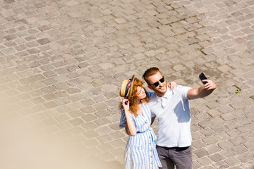 high angle view of smiling redhead man in sunglasses taking selfie with girlfriend on smartphone at city street