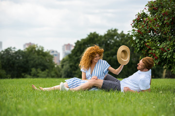 smiling redhead woman trying to putting on own straw hat on boyfriend head on grass in park