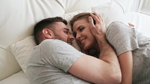 Handheld shot of happy bearded man and beautiful young woman lying on bed and looking each other in eyes