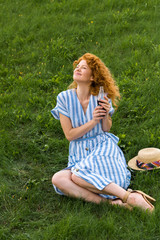 Fototapeta premium smiling redhead woman sitting on grassy meadow with soda