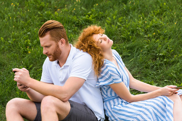 redhead couple with smartphones sitting back to back on grassy meadow