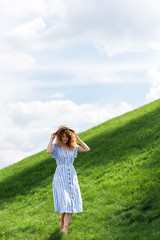 Obraz premium beautiful redhead woman in straw hat walking on grassy hill with blue sky behind