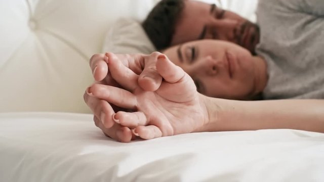 Handheld PAN of bearded man lying with closed eyes in bed and hugging beautiful young woman: they are holding hands and caressing each others fingers while enjoying intimate moment in morning