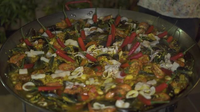 Cooking spanish paella with red hot peppers and fresh seafood in pan close up. Preparation traditional spain food paella with mussels, shrimps, calamari and vegetables in pan.