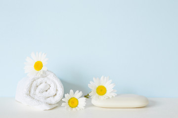 Fototapeta na wymiar White natural soap and towel on shelf in bathroom. Beautiful camomiles or daisies. Fresh flowers. Care about clean and soft face, hands, legs and body skin. Empty place for text on pastel blue wall.