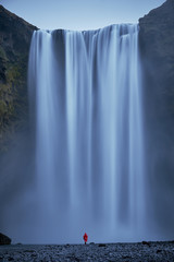 Tourist with red jacket in front of Skogafoss waterfall in Iceland island during an outdoor vacation hike in the pure wild nature 