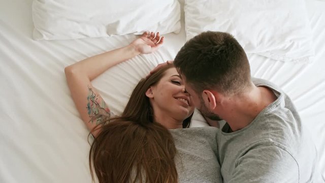 Handheld top view shot of happy young woman lying down on soft bed and laughing as her bearded boyfriend kissing her on lips