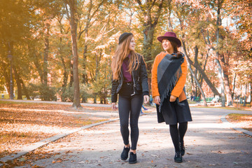 Fototapeta premium Two teenage girls are having fun in the park, autumn outfit. Funny girl friends throwing dry leaves in the city in autumn. Happy family on autumn walk!