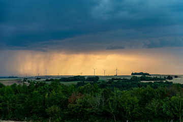 Paysage de campagne au crépuscule orange, l'orage se prépare à éclater, la pluie au loin lazarde le ciel au dessus de l'horizon et des éoliennes