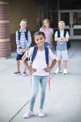 Photo of a diverse group of smiling elementary school students standing in front of the school building. Selective focus on the cute girl in front. Standing out from the crowd