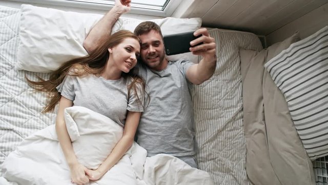 Handheld top view shot of happy young woman and bearded man lying in bed and taking selfie on mobile phone
