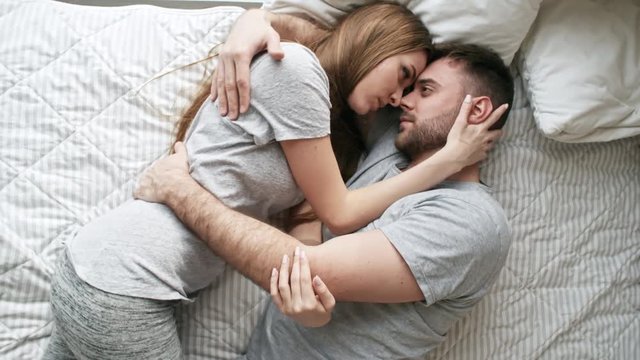 Handheld top view of affectionate young woman and bearded man lying on bed and hugging: they are looking into each others eyes and sharing intimate moment