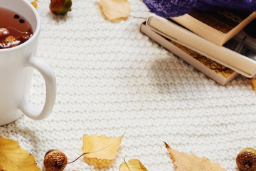 Fall composition. Cup of hot berry tea, yellow autumn leaves, acorns, warm hat and pile of books on white knitted background