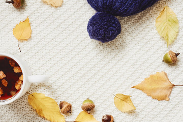 Fall composition. Cup of hot berry tea, yellow autumn leaves, acorns, warm hat on white knitted background