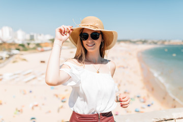 Happy smiling attractive woman on summer vacation on a seafront promenade smile at the camera in a big straw hat and sunglasses.