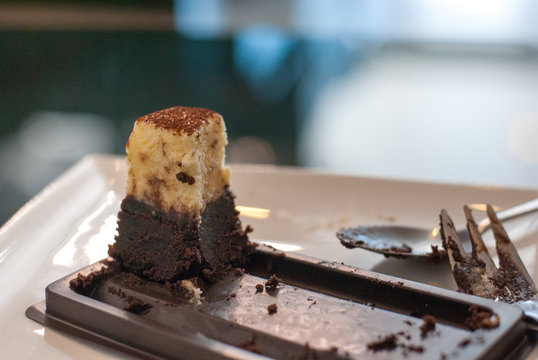 The Last Bite Of Tasty Cheese Cake Left On White Plate With Fork And Spoon Beside In Indoor Light, The Cake Itself Is Half Chocolate Half Cheese