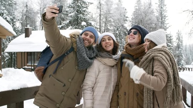 Young happy friends standing in the park at cold winter day, smiling and posing at smartphone camera while taking selfie together