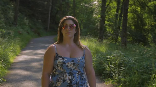 Woman in sunglasses and blue dress walking towards camera on narrow path through forest with sun flare