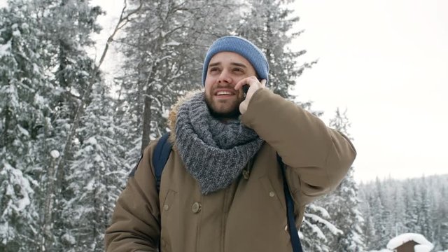 Handsome male tourist smiling and speaking on mobile phone while standing outdoors in forest at snowy winter day