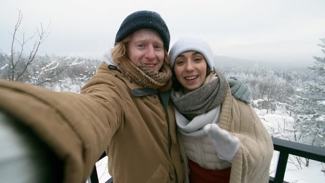 Young happy boyfriend and girlfriend standing at viewing point in national park at cold winter day, smiling and waving at camera while chatting via video call