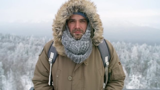 Young man with icy beard standing at viewing point in national park and looking at camera at cold winter day outdoors