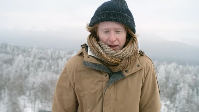 Portrait of young man standing at viewing point in national park, shivering with cold and looking at camera at frosty winter day outdoors