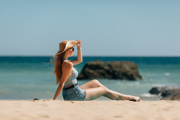 Fototapeta na wymiar Side view an attractive young woman wearing straw hat and sunglesses sits on a beach with ocean background
