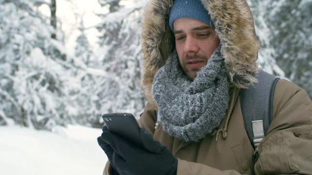 Male tourist standing in forest at frosty winter day and typing message on smartphone