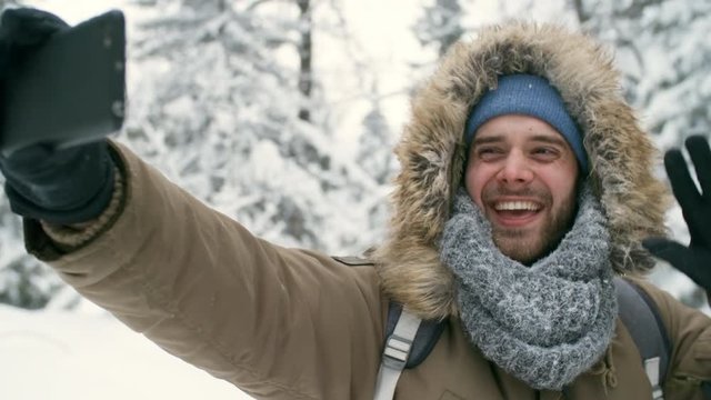 Joyful young man smiling and posing at front camera of smartphone while taking selfie in snowy forest during winter hike