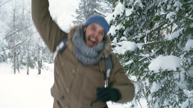 Excited man jumping up and making snow falling down from tree branch while having fun outdoors at winter day