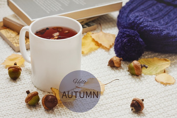 Fall composition with sing hello autumn. Cup of hot berry tea, yellow autumn leaves, acorns, warm hat and pile of books on white knitted background