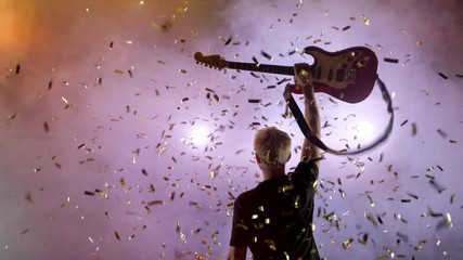 GUITARIST ON SCENE raises the guitar up, spectators applaud. The guitarist performs on stage. Stage light, smoke. From above fall golden confetti,