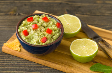 Guacamole in blue bowl on natural rustic desk with ingredient: lemon, tomatoes, peppers around.