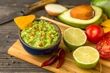 Guacamole in blue bowl on natural rustic desk with ingredient: lemon, tomatoes, peppers around.