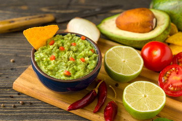 Guacamole in blue bowl on natural rustic desk with ingredient: lemon, tomatoes, peppers around.