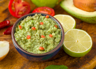 Guacamole in blue bowl on natural rustic desk with ingredient: lemon, tomatoes, peppers around.