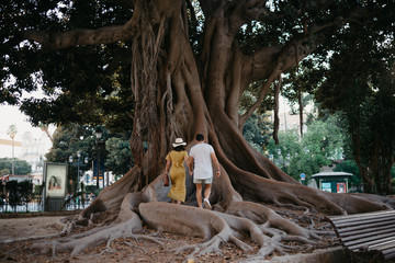 Beautiful young brunette woman with her boyfriend with beard walking under the giant tree with large roots in Spain in the evening