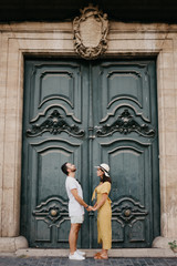 Beautiful young brunette woman with her boyfriend with beard staying holding hands with an old giant historic door on the background in Spain in the evening