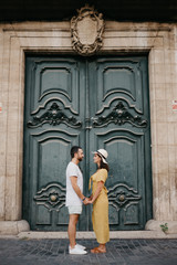 Beautiful brunette girl with her boyfriend with beard staying holding hands with an old giant historic door on the background in Spain in the evening