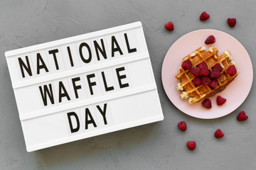 Lightbox with text 'National Waffle Day' and traditional belgian waffle with raspberries on pink plate over gray background, top view.