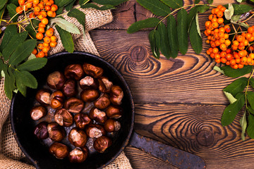 Autumn background with roasted chestnuts in a frying pan and rowan branches on a wooden table