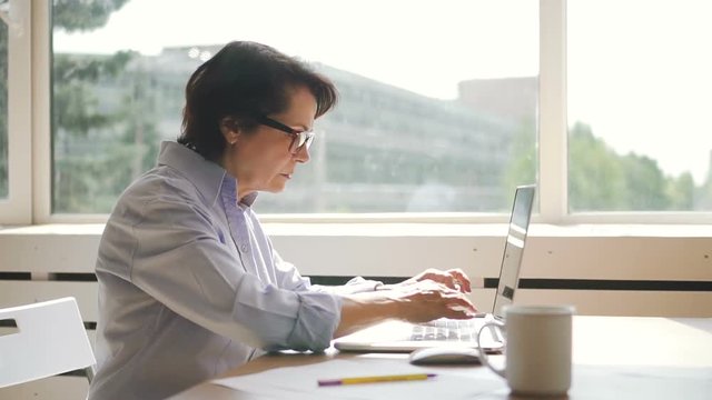 Side view of elderly businesswoman is working with laptop in home workplace, busy business person is typing, looking at pc screen, sitting at table by window in modern dwelling. Concept: mature female