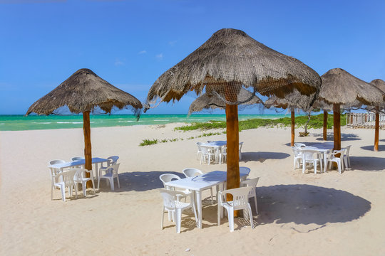 Beach bar - Tiki thatch umbrellas on sandy beach with white plastic chairs underneath and the turquioise sea and very blue sky on background