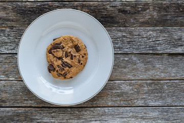 Single chocolate chip cookie on a round white plate sitting on weathered barn wood background.