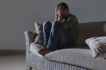 lifestyle dramatic light portrait of young sad and depressed man sitting at shady home couch in pain and depression feeling stressed and desperate