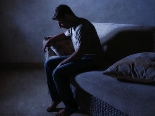 lifestyle dramatic light portrait of young sad and depressed man sitting at shady home couch in pain and depression feeling stressed and desperate